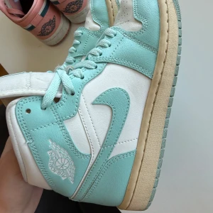 Nike Air Jordan 1 Mintgrön/Vit - Säljer ett par Nike Air Jordan 1 i mintgrönt och vitt skinn med klassisk Jordan-logga på sidan. Skorna har snörning, rund tå och en retro beige sula som ger en snygg kontrast. Perfekta för dig som vill sticka ut med stilrena sneakers.