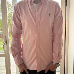Ljusrosa skjorta från Ralph Lauren - Säljer en ljusrosa skjorta från Ralph Lauren i slim fit med klassisk button down-krage och broderad logga på bröstet. Skjortan är långärmad, har vita knappar och är tillverkad i mjuk bomull. Perfekt för dig som gillar stilrena och snygga plagg.