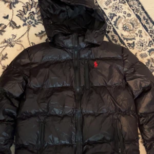 Svart pufferjacka från Polo Ralph Lauren XL - Svart pufferjacka från Polo Ralph Lauren i storlek XL. Jackan har huva, flera dragkedjor framtill och det klassiska röda Polo-logot på bröstet. Materialet är glansigt och syntetiskt, perfekt för kalla dagar. Jackan är långärmad och har en loose passform.