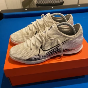 Nike vita fotbollsskor med mönster - Säljer ett par vita Nike fotbollsskor med svart Swoosh och coolt grafiskt mönster på sidan. Skorna har snörning, platt sula och är tillverkade i syntetmaterial med mesh för bra andningsförmåga. Perfekta för inomhusspel och har en modern, sportig look. Priset är inte hugget i sten, inga skambud