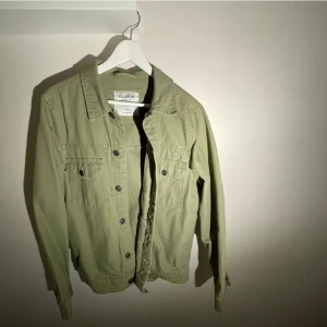 Olivgrön jeansjacka från Pull&Bear - Säljer en olivgrön jeansjacka från Pull&Bear i storlek S. Jackan har klassisk krage, två bröstfickor med knappar och knäppning framtill. Tillverkad i bomull och har en rak, normal passform. Perfekt för lager-på-lager och enkel att styla.