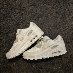 Nike Air Max 90 vita sneakers - Nike Air Max 90 sneakers i vitt med ljusgrå och beige detaljer. Klassisk siluett med synlig Air Max-dämpning i sulan, mesh och skinnpaneler samt snörning. Swoosh-logga i beige och grönaktig detalj på hälen. Perfekta för dig som gillar stilrena och fräscha sneakers.