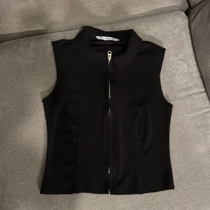 Svart ärmlös topp med dragkedja från MNG - Snygg svart ärmlös topp från MNG med krage och silverfärgad dragkedja framtill. Toppen har en figurnära passform och stilren design som passar perfekt till jeans eller kjol. Materialet känns mjukt och följsamt.