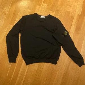 Svart sweatshirt från Stone Island - Svart sweatshirt från Stone Island med klassisk rund hals och ribbade muddar. Ikonisk logotyp-patch på vänster ärm. Tröjan är i mjuk bomull och har en clean, stilren look som passar perfekt till streetwear-stilen.