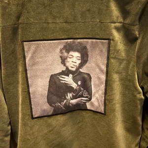 Olivgrön bomberjacka från Limitato - Snygg olivgrön bomberjacka från Limitato med mjuk sammetkänsla och guldfärgade dragkedjor. På ryggen finns en stor svartvit patch med konstnärligt foto. Jackan har ribbad krage och två fickor med dragkedja framtill.