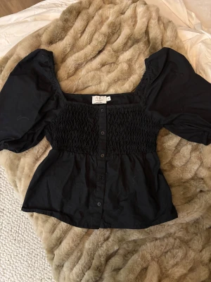 Svart blus med puffärm från AFJ x NA-KD - Supersnygg svart blus med puffiga korta ärmar och smockad överdel från AFJ x NA-KD. Blusen har knappar framtill och fyrkantig ringning som ger en trendig vibe. Tillverkad i bomull, perfekt att styla med jeans eller kjol för en cool look.