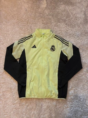 Real Madrid vindjacka från Adidas - Gul och svart vindjacka från Adidas med Real Madrid-logga på bröstet och klassiska Adidas-ränder på axlarna. Jackan har hel dragkedja, långärmade svarta ärmar och lätt material som passar perfekt för fotbollsträning. Snygg sportig look för dig som gillar Real Madrid.