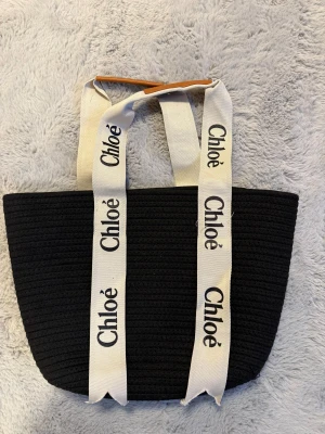 Chloé Woody Tote Bag – perfekt present till flickvän eller mamma! - Säljer en stilren och snygg Chloé Woody Tote i svart med klassiska beige band med Chloé-logo. Väskan är rymlig, lätt och passar både till vardag och fest. Perfekt att ge bort som present – uppskattas alltid av både flickvänner, mammor och systrar.  Skick: Fint begagnat skick Färg: Svart med beige handtag Detaljer: Ikoniska Chloé-band, mjukt och hållbart material Användning: Funkar super till shopping, jobb, skola eller weekendresor  Perfekt för någon som gillar lyxiga och stilrena accessoarer