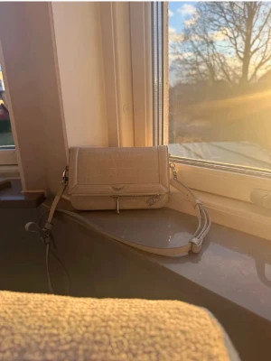 Zardig voltaire - Snygg beige axelväska med croco-mönstrad yta och justerbar rem. Väskan har ett lock med magnetstängning och en liten dragkedjeficka framtill. Perfekt storlek för mobil och småsaker, stilren och trendig design.