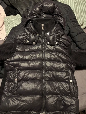 Svart Moncler pufferjacka med huva - Svart pufferjacka från Moncler med stickade svarta ärmar och huva. Jackan har dragkedja framtill, tryckknappar och två fickor med logodetaljer. Insidan har en unik serietidningsetikett. Passar S