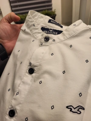 Muscle fit vit henley-skjorta från Hollister S - Säljer en vit henley-skjorta från Hollister med svarta knappar och små svarta diamantmönster. Skjortan har muscle fit-passform, lång ärm och klassisk Hollister-logga broderad på bröstet. Materialet är stretchigt bomullstyg för extra komfort.