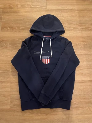 Mörkblå hoodie från GANT med tryck - Snygg mörkblå hoodie från GANT med vit dragsko i huvan och broderad logga samt sköld med stjärnor och ränder på bröstet. Klassisk känguruficka framtill. Mjuk bomullsblandning som är skön att ha på sig.