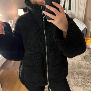 Teddyjacka svart med dragkedja - Mysig svart teddyjacka med hög krage och grov dragkedja framtill. Jackan har två stora fickor och en oversized passform som ger en chill vibe. Perfekt för kalla dagar när du vill vara både varm och snygg. Materialet är fluffigt och mjukt, vilket gör den extra skön att bära.