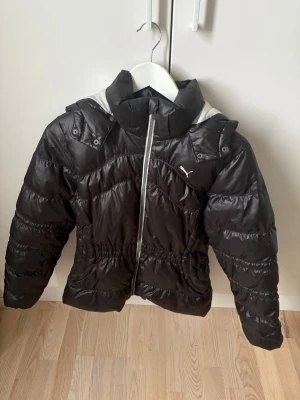Svart pufferjacka från Puma, M - Svart pufferjacka från Puma med vit kontrast på huvan. Jackan har en snygg quiltad design, dragkedja framtill och elastisk midja för skön passform. Perfekt för kalla dagar och har en sportig vibe med Puma-loggan på bröstet. Det ända är att de är ett litet hål över storlekslappen som lätt kan fixas. 