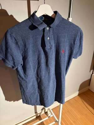 Blå pikétröja Polo Ralph Lauren M - Klassisk blå pikétröja från Polo Ralph Lauren i slim fit. Tröjan har korta ärmar, krage, två knappar och den ikoniska röda loggan broderad på bröstet. Perfekt för en clean och stilren look.