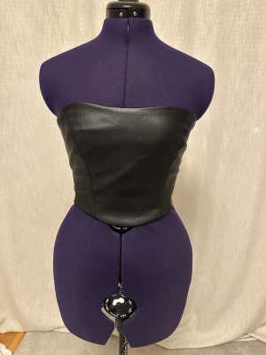 Svart skinnkorsett från Nelly - Säljer en svart axelbandslös korsett från Nelly i skinnimitation. Toppen är tight och har dragkedja bak för enkel på- och avtagning. Perfekt för att skapa en edgy look och passar till både jeans och kjol.