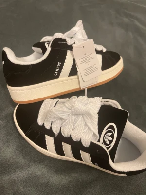 Adidas Campus sneakers svart/vit - Adidas Campus sneakers i svart mocka med vita detaljer och klassiska tre ränder på sidan. Skorna har rund tå, platt sula och breda vita snören. Ikonisk Adidas-logga på plösen och Campus-tryck på sidan. Perfekt för dig som gillar retrostil och streetwear.