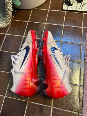 Nike Mercurial Air Zoom fotbollsskor - Säljer ett par Nike Mercurial Air Zoom fotbollsskor i vitt och rött med svarta detaljer. Skorna har snörning, platt sula med dobbar och en snygg gradient från vitt till rött. Perfekta för dig som vill sticka ut på planen med stilrena och lätta skor.