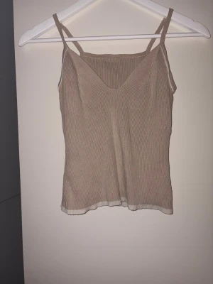 Beige ribbad linnetopp med smala band - Säljer en beige ribbad linnetopp med smala axelband och v-ringning framtill. Toppen har en lätt croppad passform och vit kant längst ner. Perfekt för en chill och stilren look.