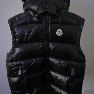 Svart dunväst från Moncler - Snygg svart dunväst från Moncler med glansig finish och ikonisk logga på bröstet. Västen har huva, dragkedja framtill och två fickor med dragkedja. Perfekt för lager-på-lager och ger en clean streetstyle-vibe.