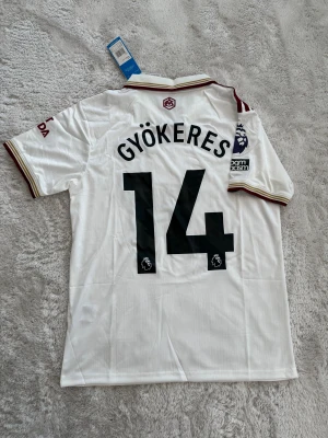Arsenal Gyökeres vit matchtröja Adidas - Säljer en fräsch Arsenal matchtröja med Gyökeres och nummer 14 på ryggen. Tröjan är vit med vinröda detaljer, klassisk krage och korta ärmar. Premier League-logga på ärmen och snygga broderade klubbmärken. Materialet är lätt polyester, perfekt för fotboll.