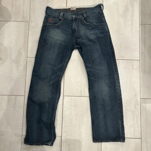 Blå jeans från Tommy Hilfiger - Snygga blå jeans från Tommy Hilfiger Denim med klassisk femficksdesign, kontrastsömmar och röd broderad logga både fram och bak. Jeansen har rak passform, tillverkade i slitstarkt denimtyg med coola detaljer på fickorna 36/32