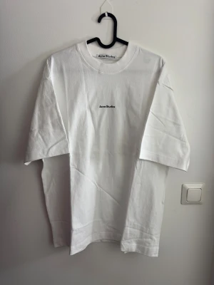 Vit t-shirt Acne Studios S - Vit t-shirt från Acne Studios i storlek S. Klassisk rund hals och korta ärmar. T-shirten är tillverkad i 100% bomull och har en stilren logotyp tryckt på bröstet. Enkel och clean design som funkar till allt. Perfekt för dig som gillar minimalistisk stil.