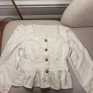 Vit blus med volang och guldknappar - Supersöt vit blus med subtilt blommönster, volang nertill och stora guldiga knappar framtill. Långärmad modell med fyrkantig ringning och lätt puff vid axlarna.