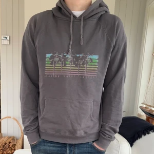 Grå hoodie Saint Laurent Paris,  Malibu hoodie - Säljer en grå hoodie från Saint Laurent med tryck av palmer och texten 'Malibu Saint Laurent' på bröstet. Tröjan har huva med snörning, känguruficka och långa ärmar. Materialet är mjuk bomull och passformen är normal. Perfekt för chill dagar.