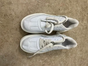 Vita Adidas sneakers i skinn - Säljer ett par klassiska vita Adidas sneakers i skinn med snörning och platt sula. Skorna har diskreta gröna och gula detaljer på häl och plös samt de ikoniska tre ränderna på sidan. Perfekta för en clean och sportig look.