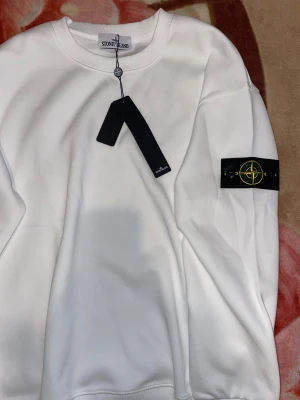 Vit Stone Island sweatshirt M - Vit sweatshirt från Stone Island med klassisk rund hals och ribbade muddar. Tröjan har den ikoniska svarta patchen med kompasslogga på vänster ärm. Tillverkad i mjuk bomull för en clean och stilren look.