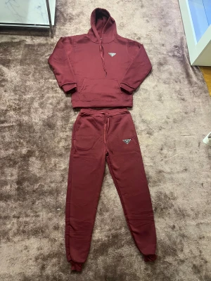 Vinröda mjukisbyxor från Prada med tröja - Snygga vinröda mjukisbyxor från Prada med klassisk logga på benet. Byxorna har normal passform, resår och snörning i midjan samt muddar vid bensluten. Materialet är mjukt och skönt, perfekt för chill dagar. Matcha med hoodien för en clean look.