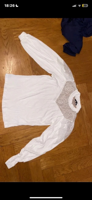 Vit spetsblus från Happy Holly - Supersöt vit blus från Happy Holly med långärmade ärmar och transparent spets upptill vid axlar och hals. Blusen har en hög krage och är tillverkad i ett lätt, ribbat tyg som ger en snygg struktur. Perfekt för dig som gillar romantiska detaljer och stilren look.