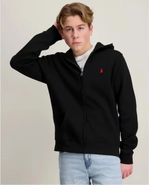 Svart hoodie från Polo Ralph Lauren - Svart hoodie från Polo Ralph Lauren med dragkedja och huva. Klassisk röd logga broderad på bröstet och känguruficka framtill. Skön passform och mjukt material, perfekt till jeans eller joggers.