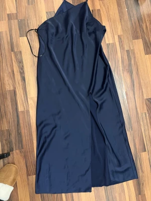 Marinblå långklänning från ASOS DESIGN - Säljer en marinblå långklänning från ASOS DESIGN med slits framtill och tunna axelband. Klänningen har en snygg omlott-look upptill och är gjord i ett glansigt, mjukt polyestermaterial som ger en lyxig känsla. Perfekt för dig som gillar stilrena och eleganta plagg.