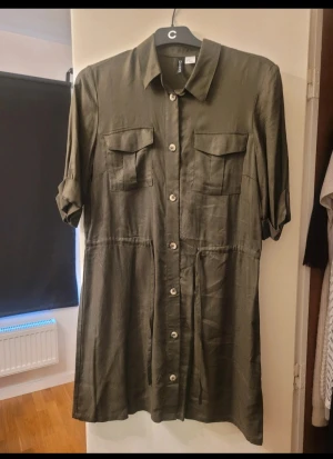 Olivgrön skjortklänning från H&M Divided - Olivgrön skjortklänning från H&M Divided i storlek 36. Klänningen har korta ärmar, knappar hela vägen framtill, två bröstfickor med lock och klassisk krage. Materialet är mjukt och har en lätt glansig finish. Perfekt för dig som gillar enkel och stilren design.