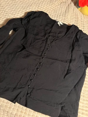 Svart långärmad blus från H&M - Svart blus från H&M i storlek S med lång ärm och snygga klädda knappar framtill. Blusen har en lätt glansig, skrynklig textur och v-ringning. Ärmsluten är smockade med knappar för extra detalj. Perfekt för dig som gillar stilrena och trendiga plagg.