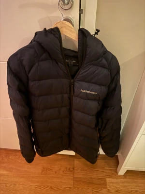 Mörkblå Peak Performance dunjacka - Säljer en mörkblå dunjacka från Peak Performance med huva och dragkedja framtill. Jackan har quiltad design och logotyp broderad på bröstet. Storleken är M på jackan men säger den passar mig som är S väldigt bra. 