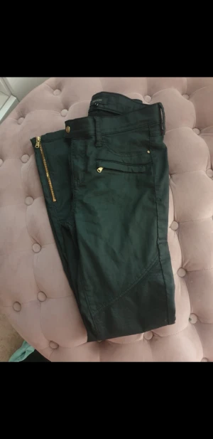 Svarta River Island byxor med gulddetaljer - Snygga svarta byxor från River Island med guldfärgade dragkedjor och knappar. Byxorna har coola sömdetaljer på benen och fickor både fram och bak. Dessa slutar vid anklarna och ser ut som skinnbyxor, sitter skit snyggt på, men dom är tyvärr för små för mig så har nog aldrig använt dessa. Finns ej att köpa längre🌸