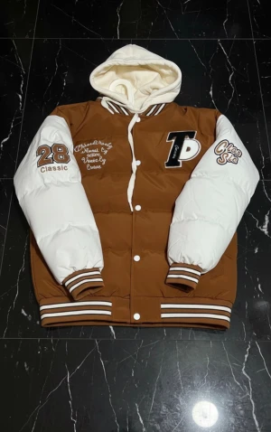 Brun och vit puffer bomberjacka med huva - Snygg brun och vit puffer bomberjacka med broderade patches och tryck på ärmar och bröst. Jackan har en mysig vit huva, ribbade muddar med ränder och knäppning framtill. Perfekt för dig som vill sticka ut med en sportig och trendig look.