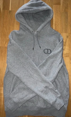 Grå hoodie från CD med CD-logga - Snygg grå hoodie från CD med broderad CD-logga på bröstet. Tröjan har huva med snörning och ribbade muddar vid ärmslut och nederkant. Mjuk insida och klassisk passform, perfekt för dig som gillar stilrena märkesplagg.