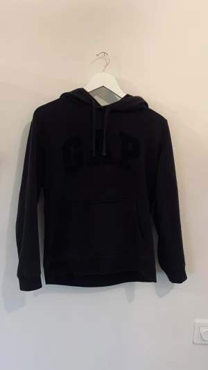 Svart hoodie från GAP XS - Svart hoodie från GAP i storlek XS med stor logga framtill. Tröjan har huva med snörning och en klassisk magficka. Mjuk insida och långärmad modell, perfekt för chill dagar. Snygg och enkel design som funkar till jeans eller mjukis.