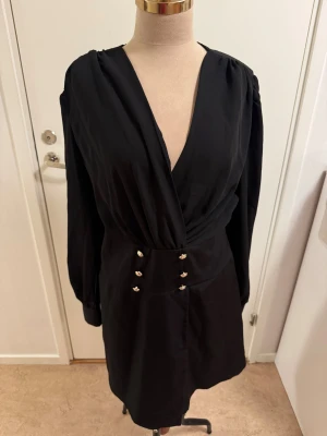 Svart omlottklänning från SHEIN XL - Svart kort omlottklänning från SHEIN med djup v-ringning, långa ärmar och dekorativa knappar i guld på midjan och ärmsluten. Klänningen har dragkedja bak och markerad midja för en snygg siluett. Perfekt för dig som gillar stilrena och trendiga plagg.