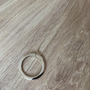 Guldigt halsband med cirkelhänge - Stilrent halsband i guldton med tunn kedja och ett stort, slätt cirkelformat hänge. Kedjan har även en liten cirkeldetalj vid låset. Perfekt för dig som gillar minimalistiska smycken med modern känsla.
