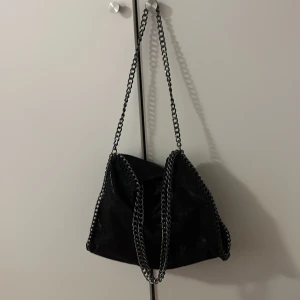 Svart axelväska med kedjedetaljer - Cool svart axelväska med glansig finish och grova kedjor längs kanterna och som axelrem. Väskan har en mjuk, slouchy form och ger en edgy vibe till din outfit. Perfekt storlek för att få plats med allt du behöver och passar till många olika stilar.