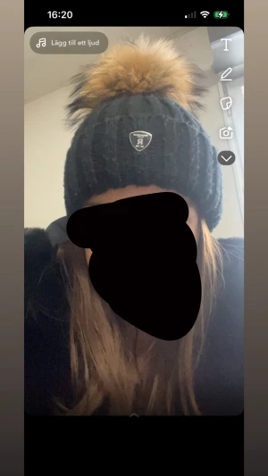 Svart stickad mössa med pälsboll - Snygg svart stickad mössa från Rock&blue med en stor fluffig pälsboll. Mössan har ett ribbat mönster och en metallbadge med logga framtill. Perfekt för kalla dagar och ger en cool vinterstil.