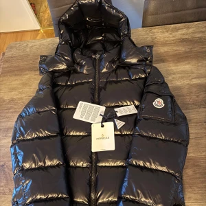 Svart pufferjacka från Moncler - Svart pufferjacka från Moncler med glansig finish, huva och klassisk logotyp på ärmen. Jackan har dragkedja framtill och quiltad design som ger en schysst volym. Perfekt för dig som vill ha en snygg och varm jacka med street vibe.