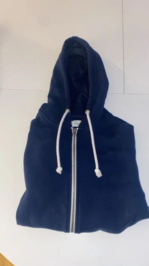 Mörkblå hoodie från Pier One - Snygg mörkblå hoodie från Pier One med dragkedja framtill och vita snören i huvan. Klassisk design med ribbade muddar och mjukt material, perfekt för chill dagar. Huvan är stor och skön, och tröjan har fickor på sidorna.