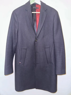 Marinblå kappa från Tommy Hilfiger - Snygg mörkblå rock från Tommy Hilfiger med klassisk krage och två framfickor. Fodret är delvis vinrött och svart, vilket ger en schysst kontrast. Jackan har en enkel knappstängning och stilren design som funkar till många looks.
