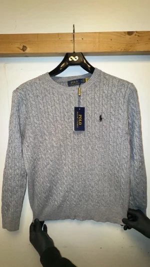 Grå kabelstickad tröja - Snygg grå kabelstickad tröja från med klassisk rund hals och diskret logga broderad på bröstet. Tröjan har långärmad passform och är tillverkad i mjuk bomull som känns skön mot huden. Perfekt för dig som gillar stilren och tidlös design.